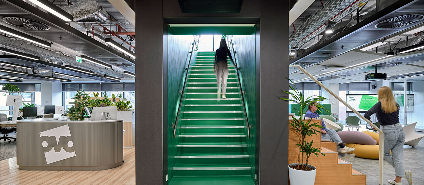 Green stairwell with man walking up. Either side of this there is a reception desk and on the other two people chatting in an office space