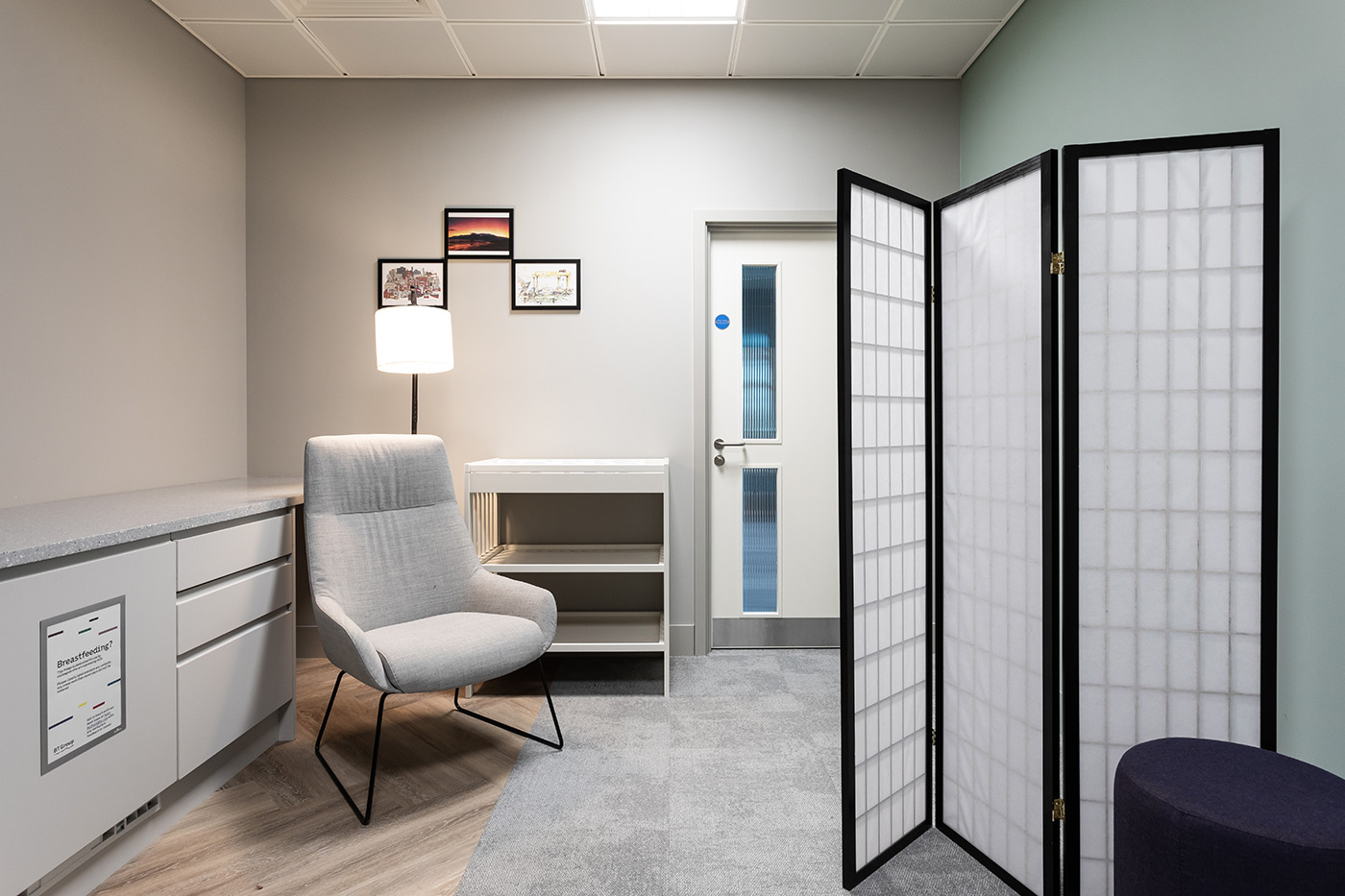 A comfortable chair beside a baby change station and a sign denoting the space for breastfeeding mothers. There is a privacy screen on the right side.